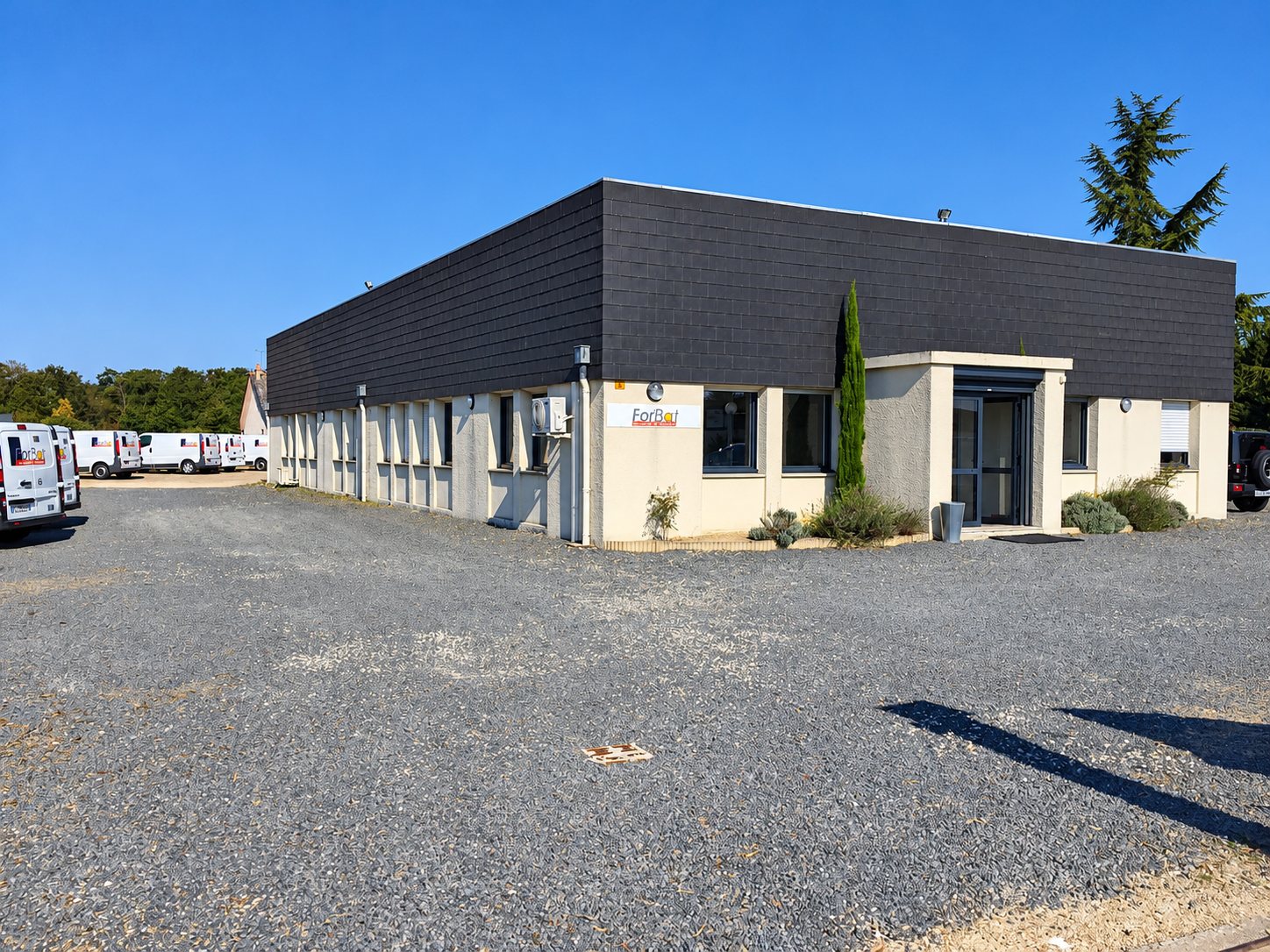 Façade d’un bâtiment de plain-pied à toit sombre portant l’enseigne ForBat, devant un grand parking gravillonné où stationnent des camionnettes blanches.
