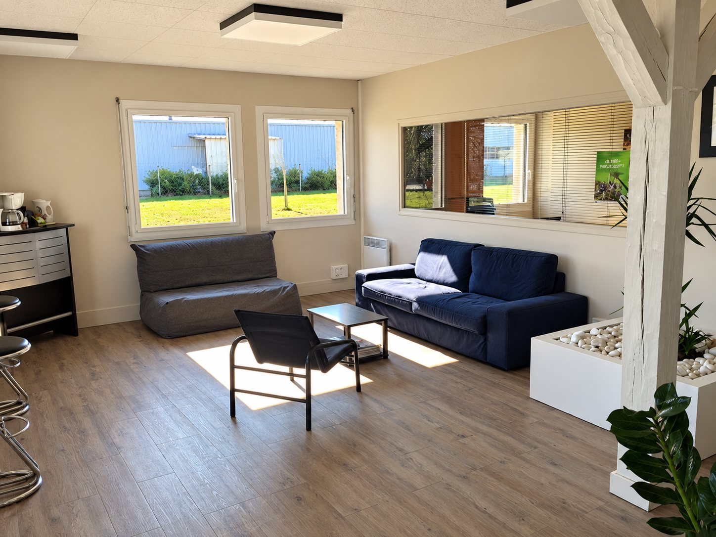 Une salle de détente lumineuse avec deux canapés sombres, un fauteuil, une table basse et des fenêtres donnant sur une pelouse.